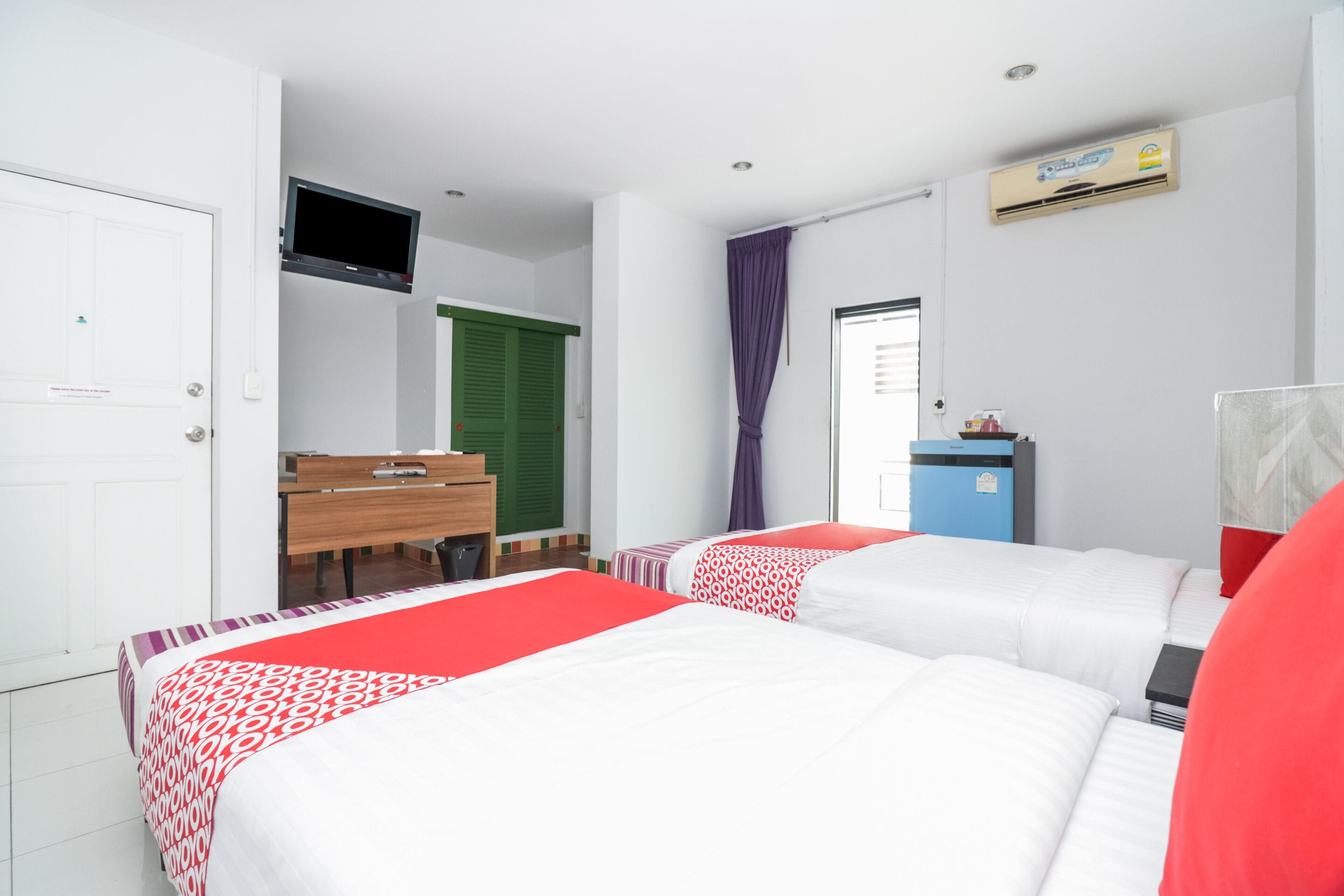 A hotel room with two beds, a desk, a television, and an air conditioning unit.