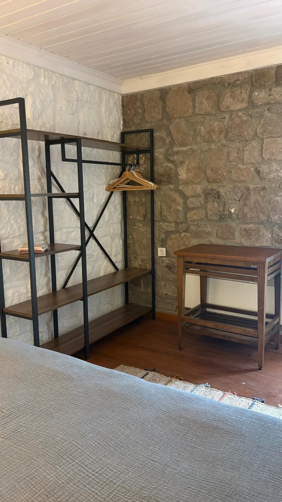 A room with a wooden floor, a black metal shelving unit with clothes hanging, and a small wooden table.