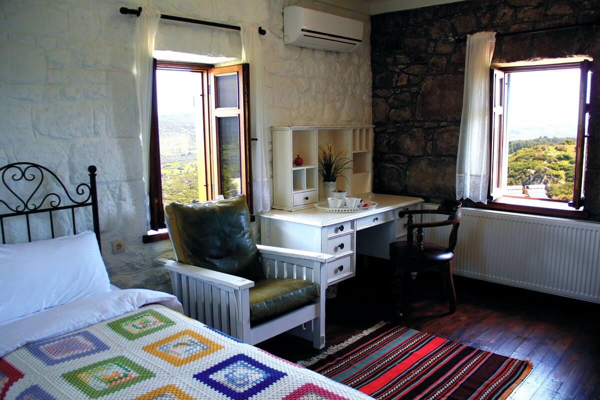 A bedroom with a bed, a desk, a chair, and a view of the outdoors.