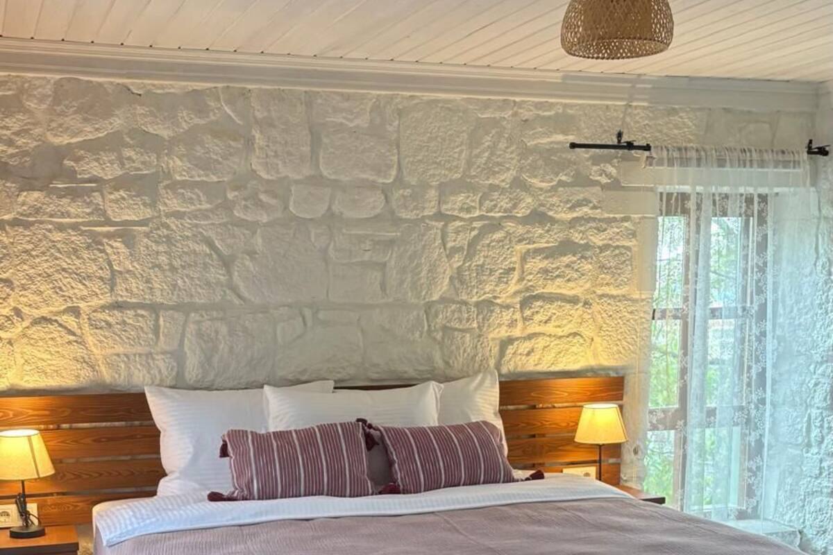 A bedroom with a stone wall, a wooden headboard, a bed with a brown blanket, two bedside lamps, and a woven rug.