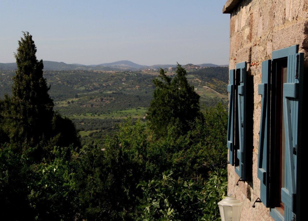 A stone building with blue shutters, overlooking a lush valley with trees and distant hills.