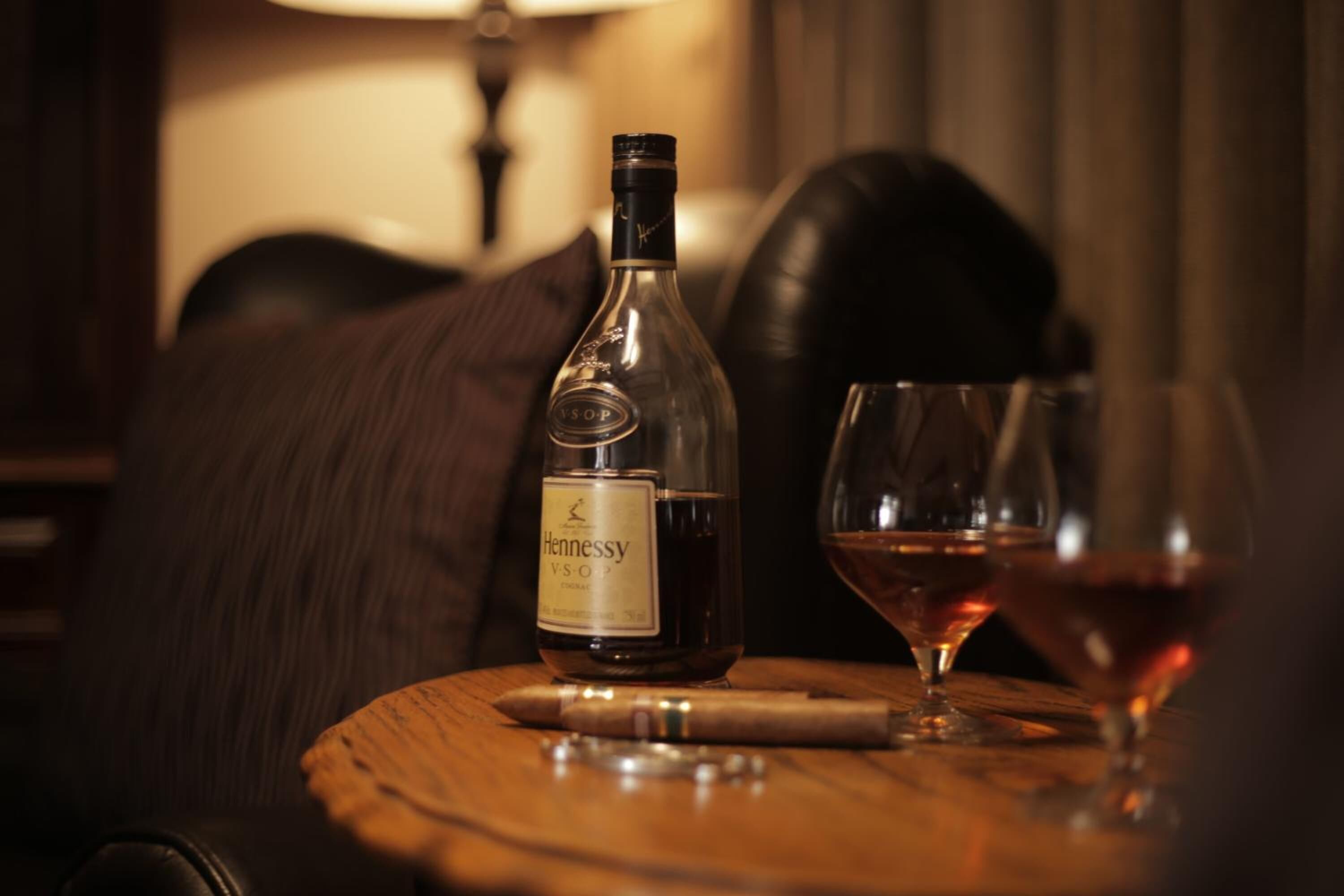 Una botella de vino Hennessy VSOP, una copa de vino y un sacacorchos sobre una mesa de madera en una habitación con poca luz.