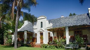 A white colonial-style building with a steeply pitched roof, a central entrance with a decorative arch, and a well-maintained garden with palm trees and benches. - Kleine Constantia (Roodepoort)