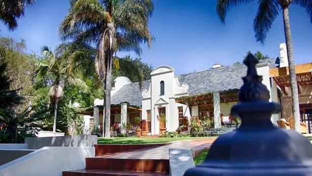 A white building with a grey roof, surrounded by palm trees and a stone pathway leading to the entrance. - Kleine Constantia (Roodepoort)
