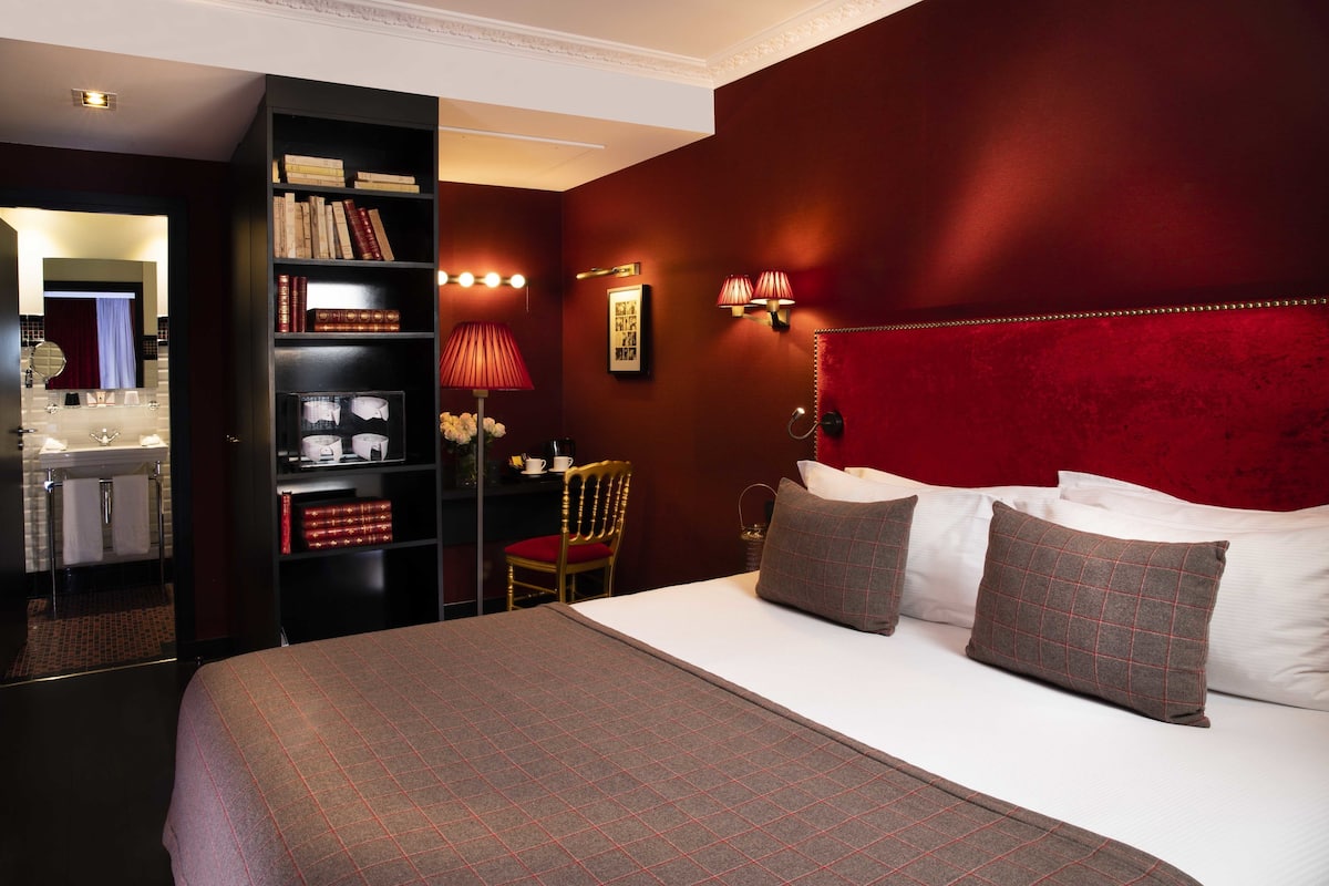 Una habitación de hotel con una cama grande, una estantería, un tocador con espejo y una pared de acento roja.