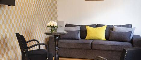 A living room with a grey sofa, two black chairs, a small round table with a vase of flowers, and patterned wallpaper.