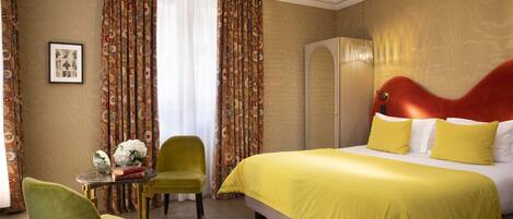 A hotel room with a bed, a green armchair, a small table with a vase of flowers, and a framed picture on the wall.