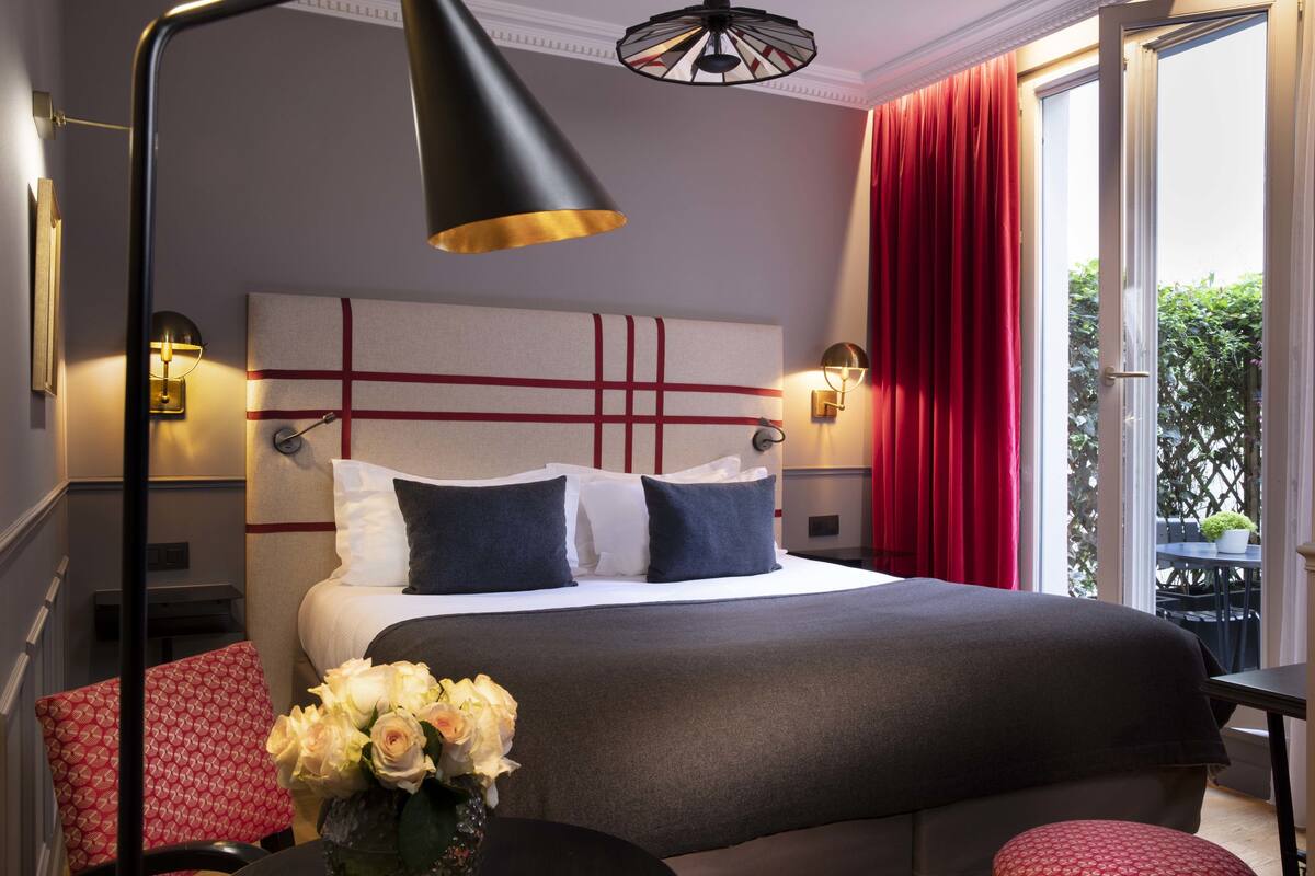 Una habitación de hotel con una cama grande, una mesita de noche, una silla y un ventanal con cortinas rojas.