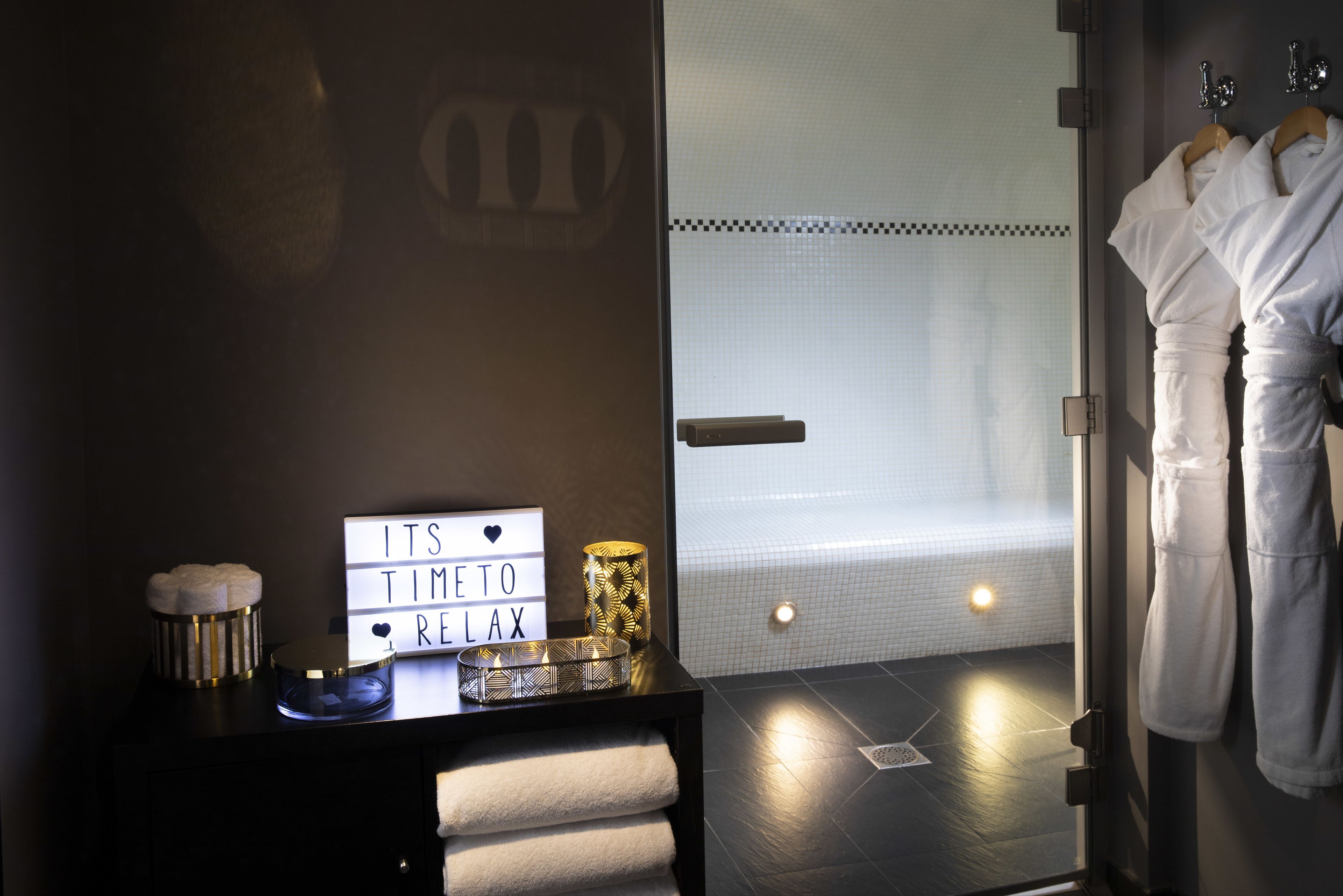 A modern bathroom with a black vanity, a light-up sign reading "IT'S TIME TO RELAX", and a shower area visible through a frosted glass door.