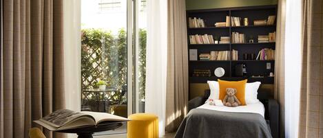 Une chambre d’hôtel avec une grande fenêtre, une bibliothèque, un lit avec un ours en peluche et un pouf jaune.