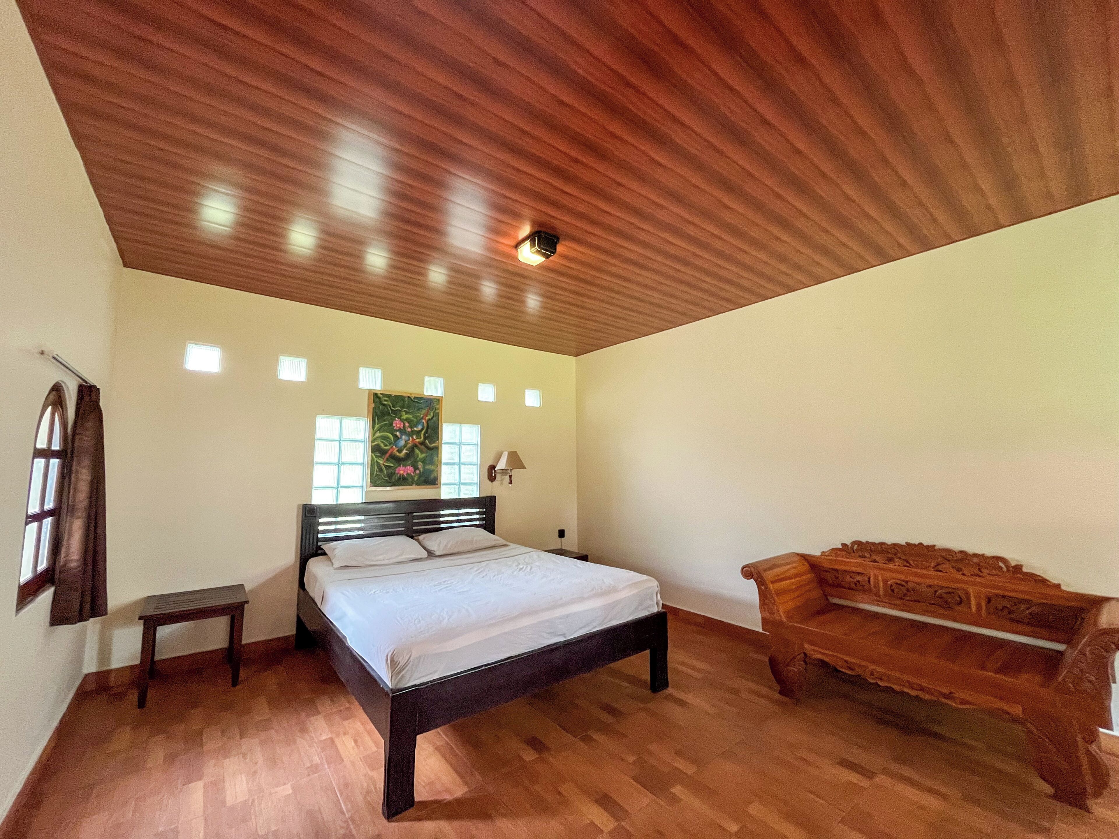 A bedroom with a wooden ceiling, a bed with white linens, a wooden bench, and a painting on the wall.