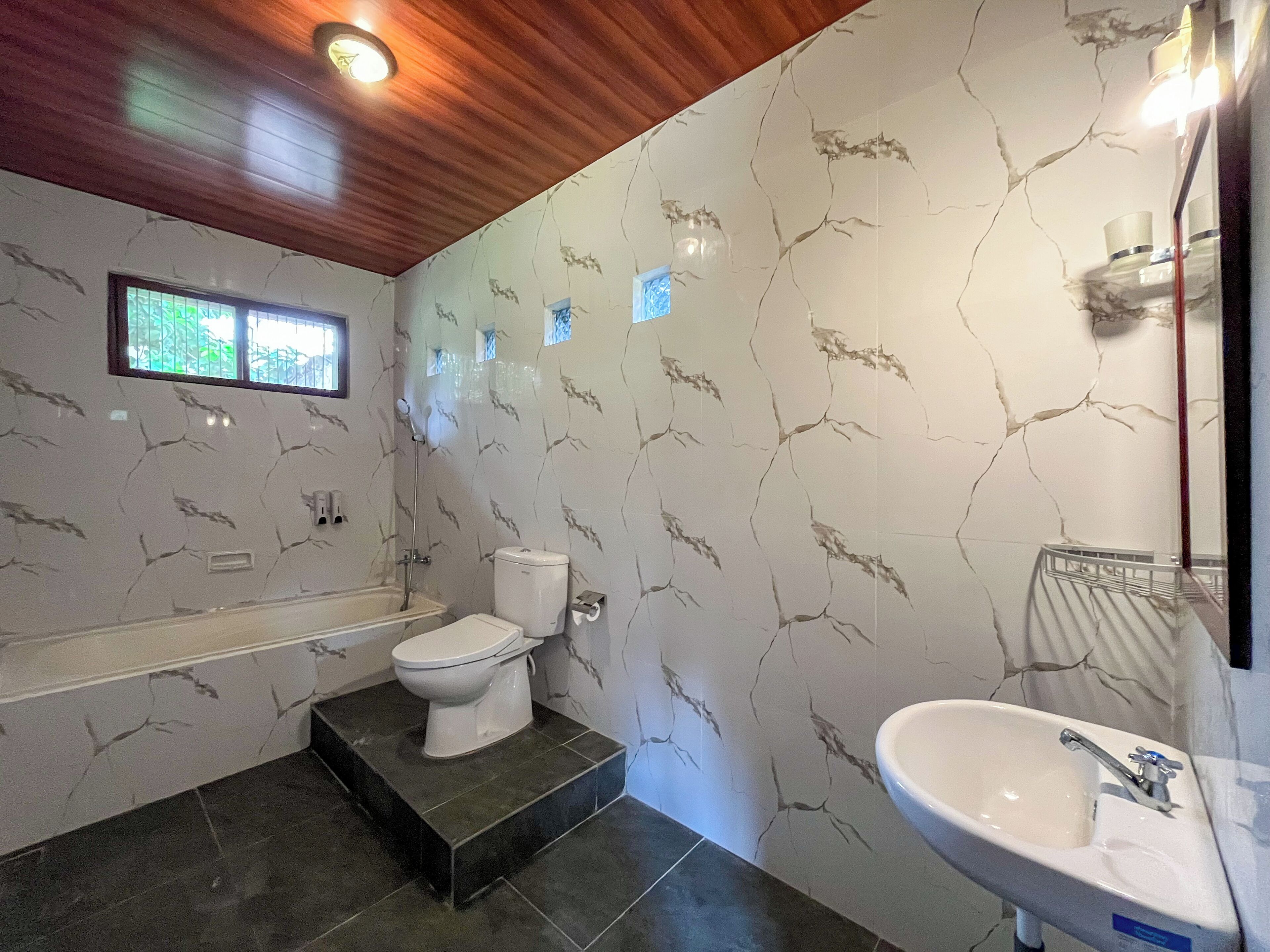 A bathroom with a bathtub, toilet, and sink, featuring marble-patterned walls and wooden ceiling.