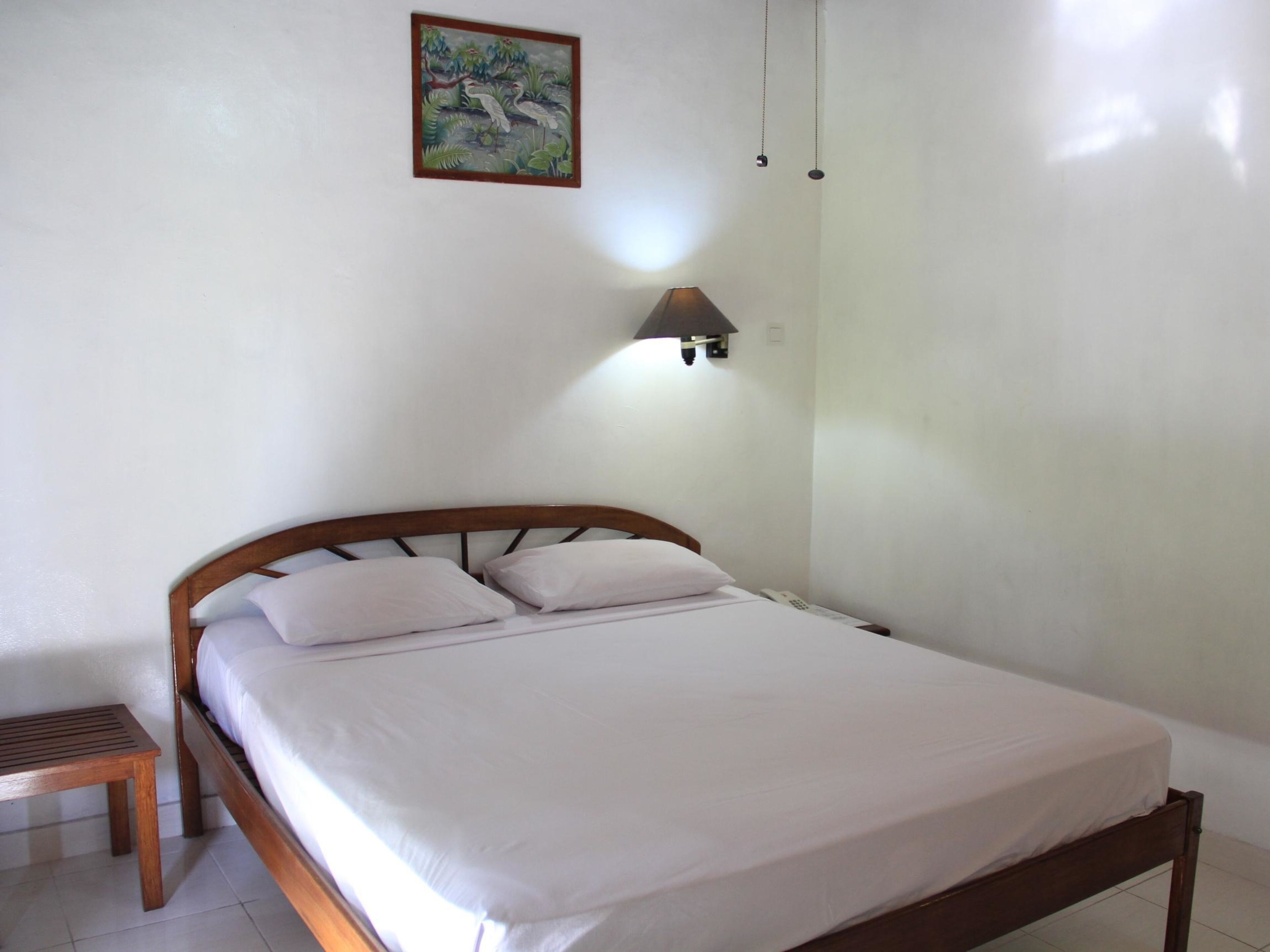 A neatly made bed with white linens in a room with white walls, a wooden headboard, a small wooden stool, and a wall-mounted light fixture.