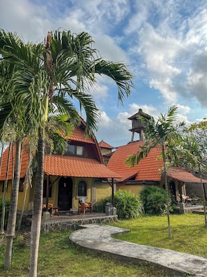 A traditional house with a tiled roof, surrounded by palm trees and a grassy area. - Suji Bungalow (Kuta)