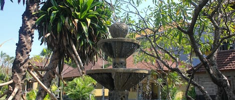 Una fuente de piedra con un adorno decorativo, rodeada de abundante vegetación y un edificio con techo de tejas al fondo.