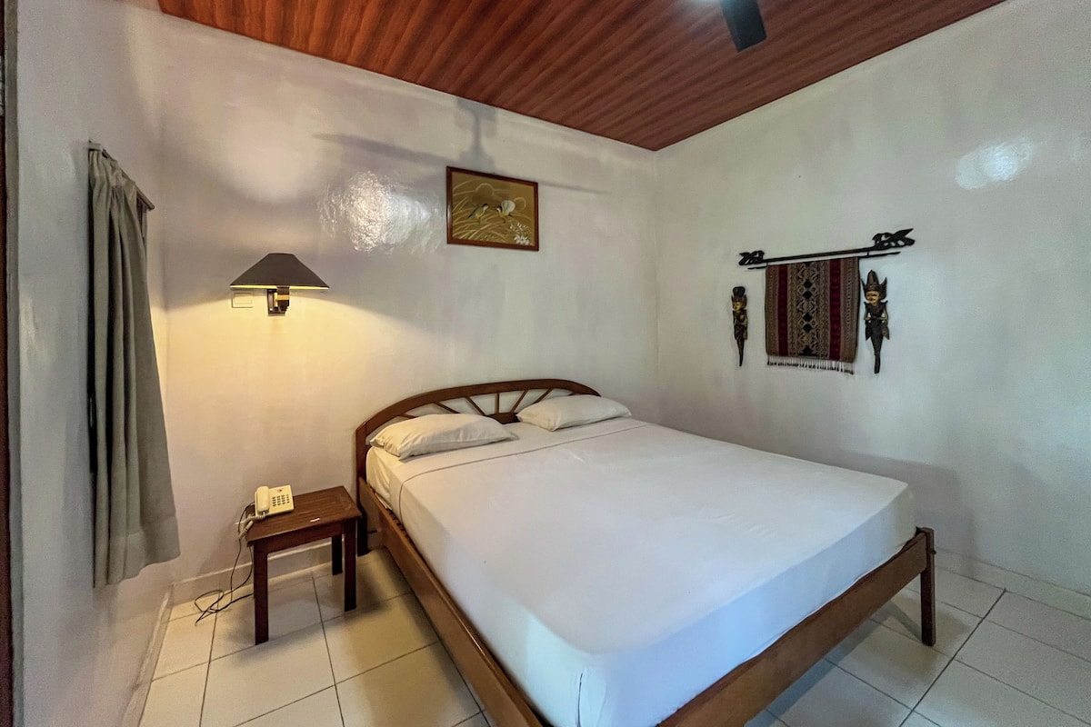 A bedroom with a wooden ceiling, a bed with white bedding, a bedside table with a lamp, and a wall-mounted decorative item.
