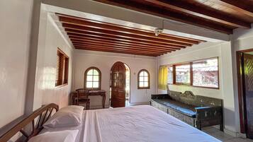 A bedroom with a large bed, wooden ceiling beams, a desk, and two arched windows.