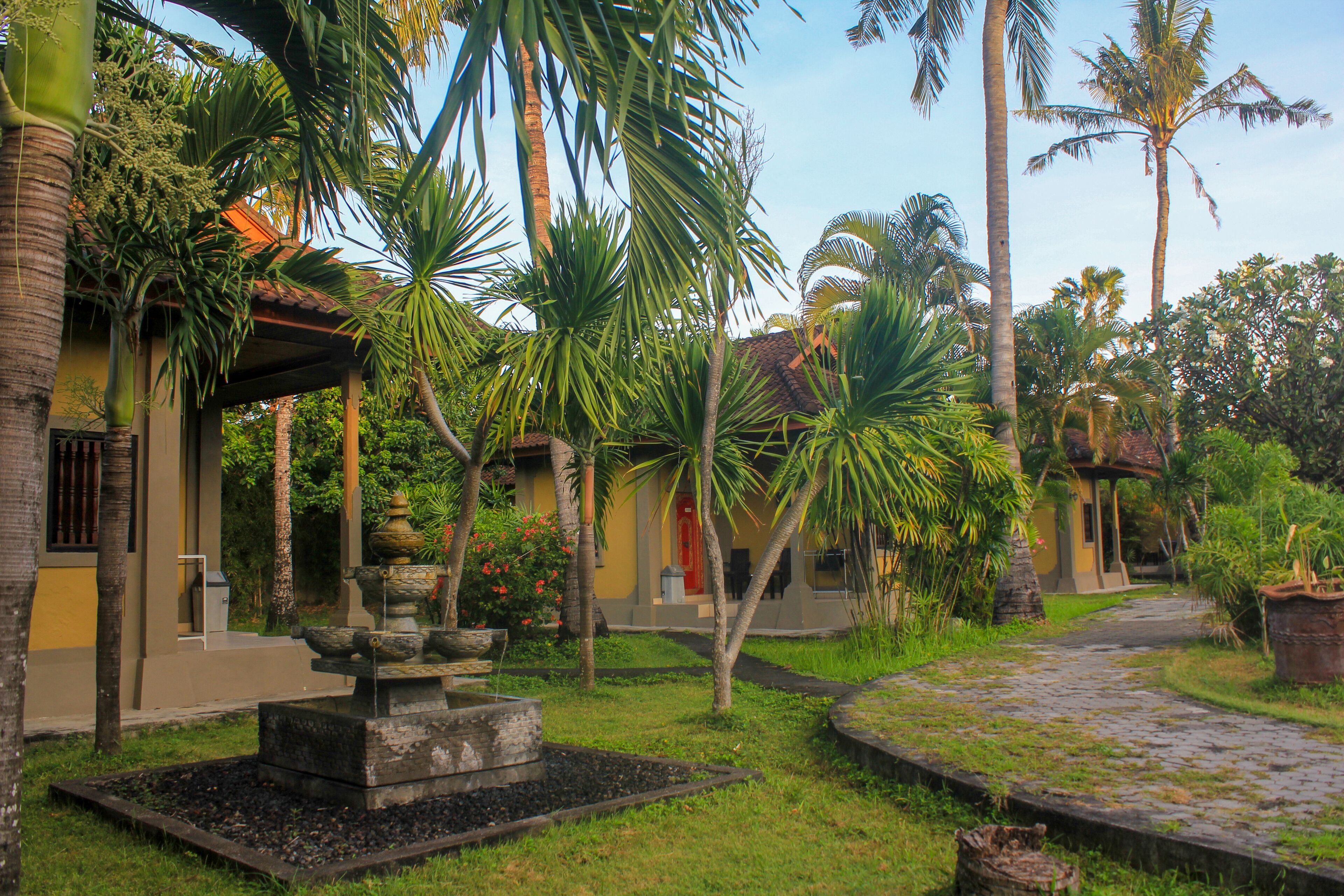 A courtyard with palm trees, a stone fountain, and a pathway leading to a building with a red door.