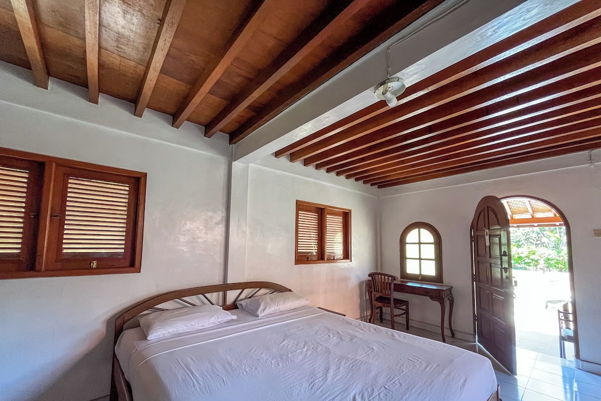 A bedroom with a bed, wooden ceiling beams, and two windows with wooden shutters.