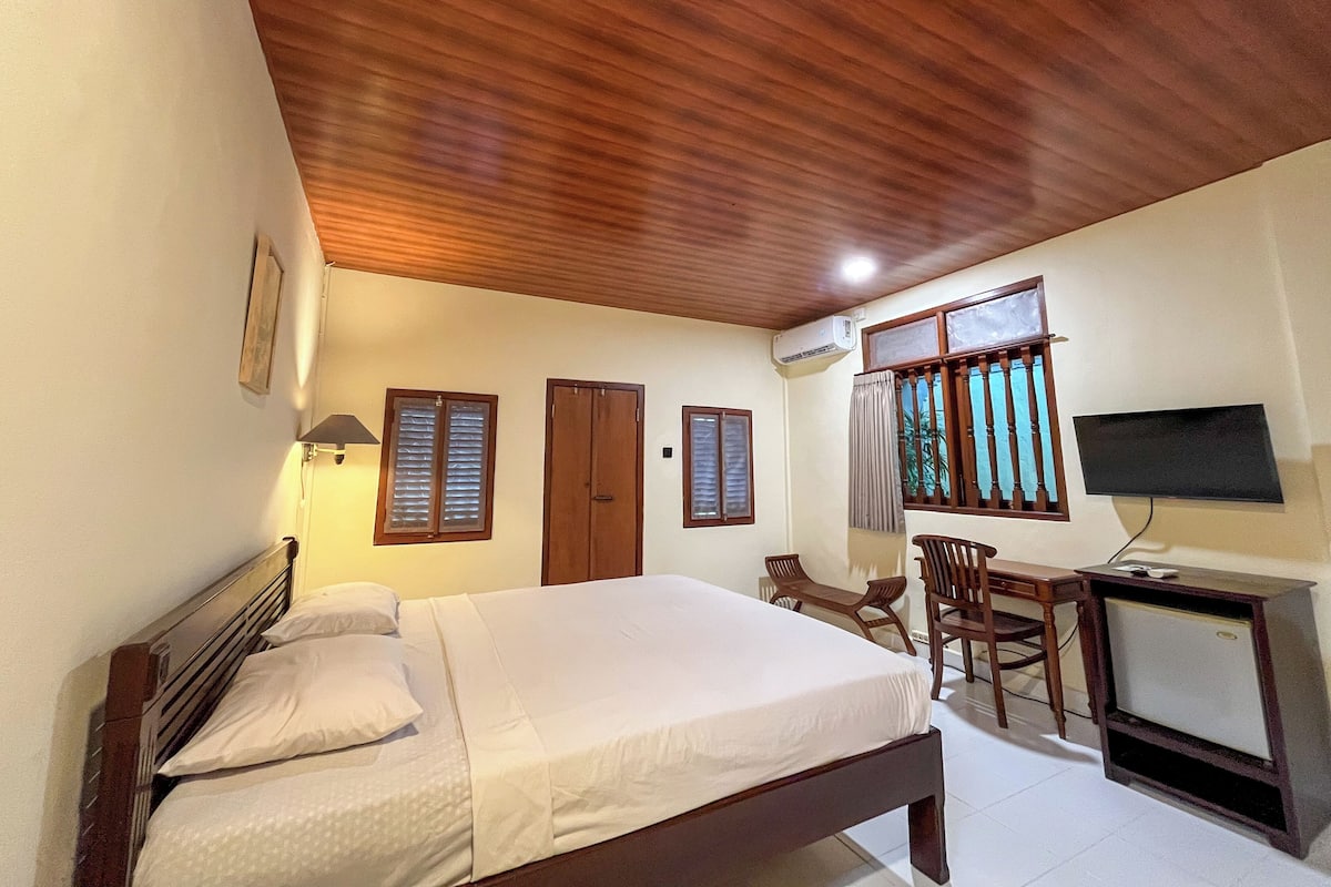 A bedroom with a wooden ceiling, a bed with white linens, a desk with a television, and two windows with shutters.
