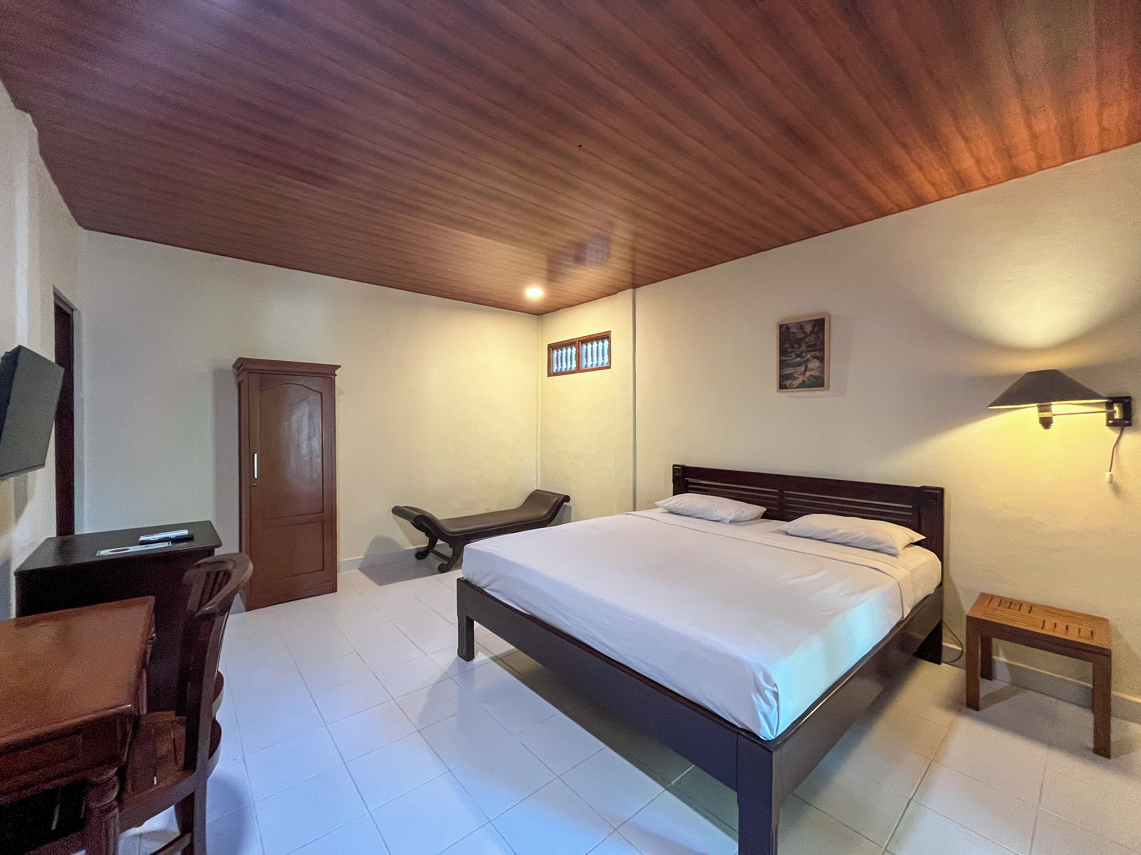 A bedroom with a wooden ceiling, a bed with white linens, a desk with a chair, a small table, and a wall-mounted lamp.