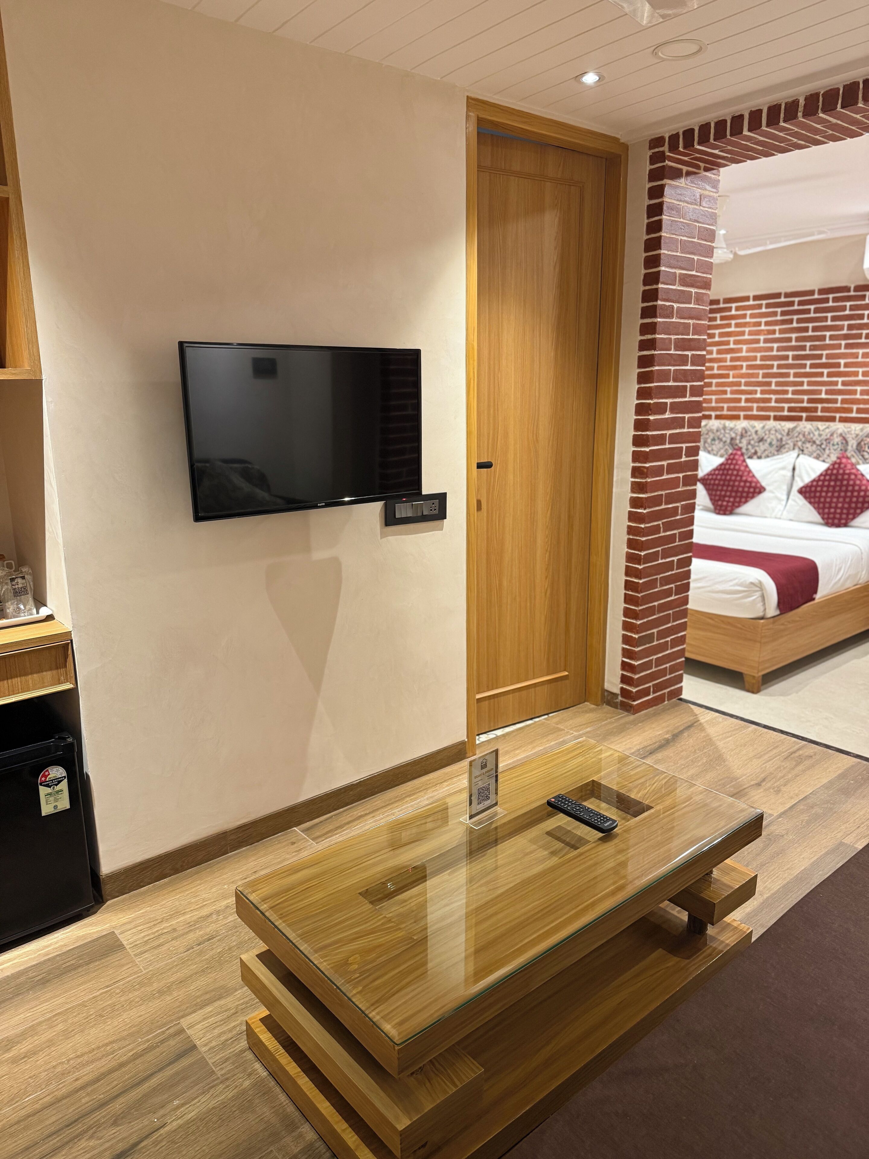 Ein Hotelzimmer mit Flachbildfernseher an der Wand, einer Holztür, einer Ziegelwand als Blickfang, einem Bett mit rot-weißer Bettwäsche und einem Holztisch mit Glasplatte, auf dem eine Fernbedienung liegt.