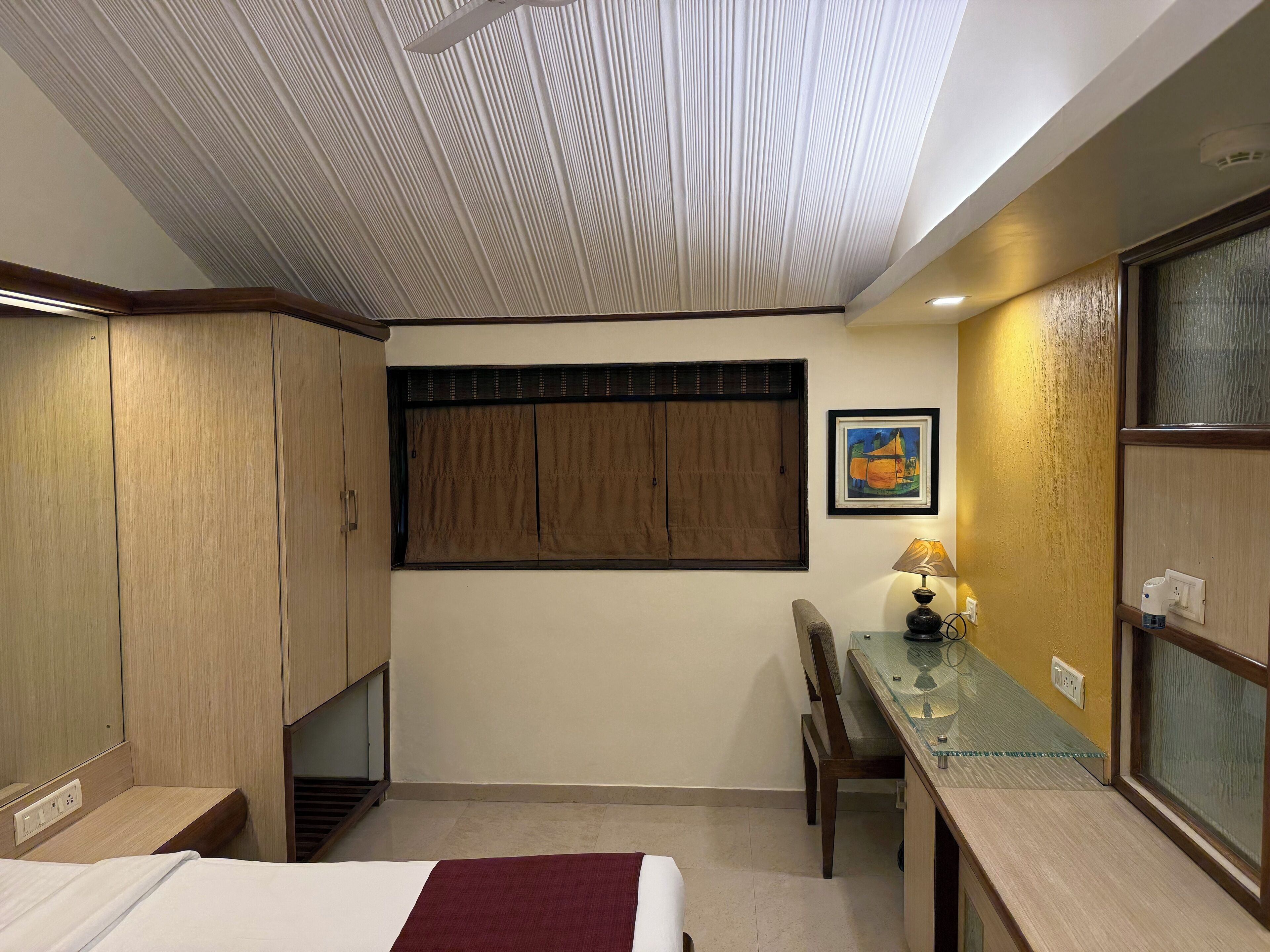 Habitación de hotel con cama, escritorio, silla, armario y techo con diseño acanalado.