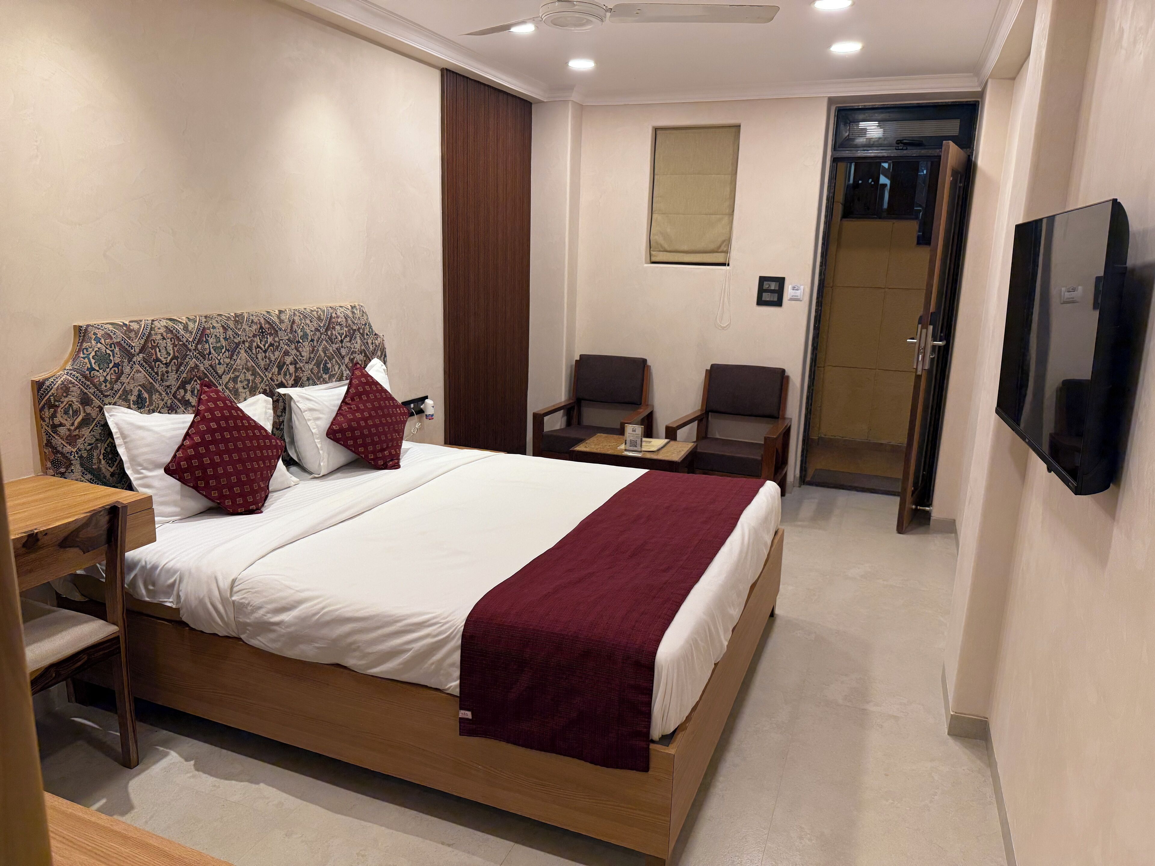 A hotel room with a large bed, a wooden nightstand, a desk with a chair, a television, and a door leading to a bathroom.