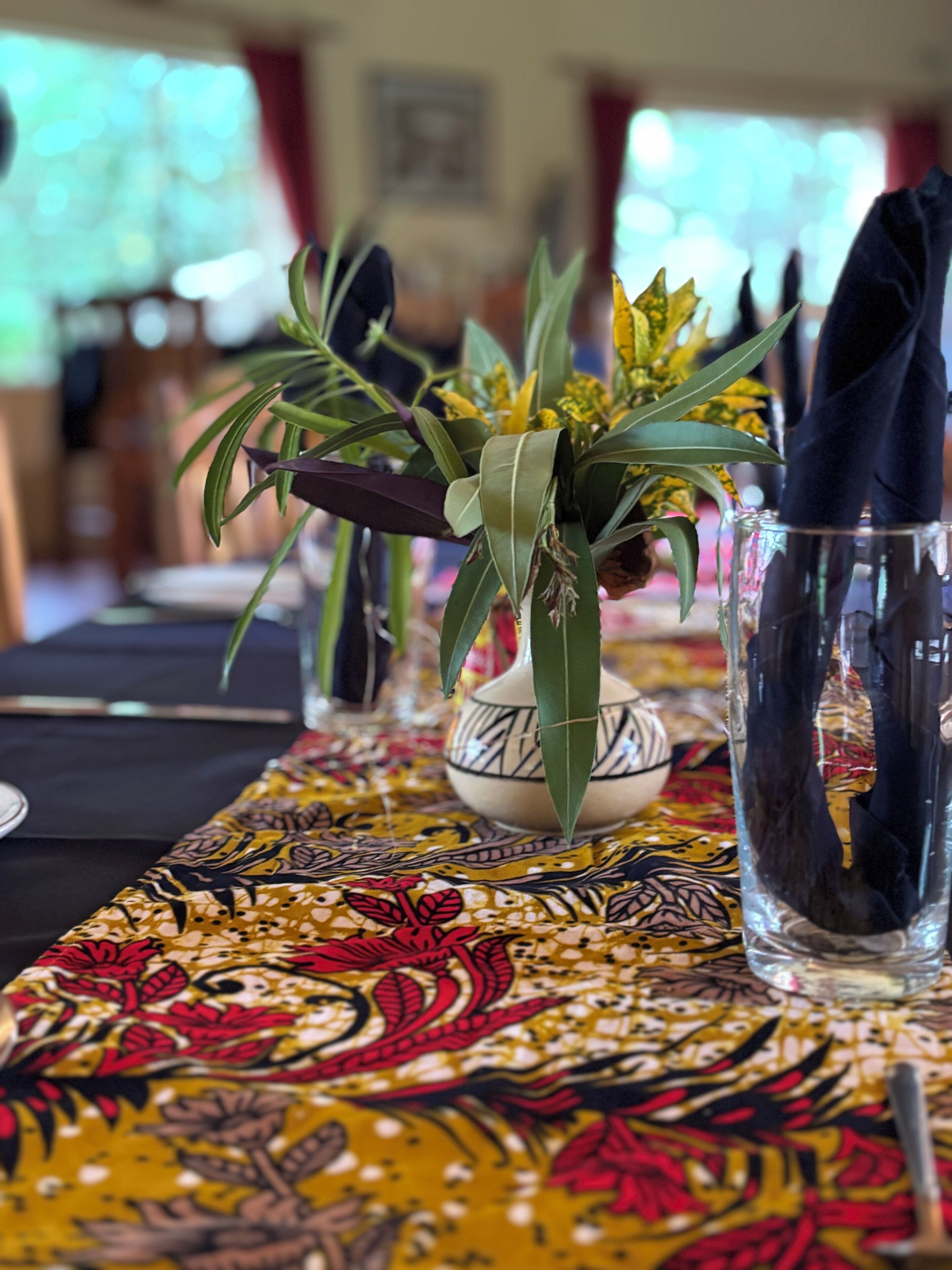 A table set with a patterned tablecloth, a vase with green leaves, a decorative bowl, and a tall glass with a dark napkin.