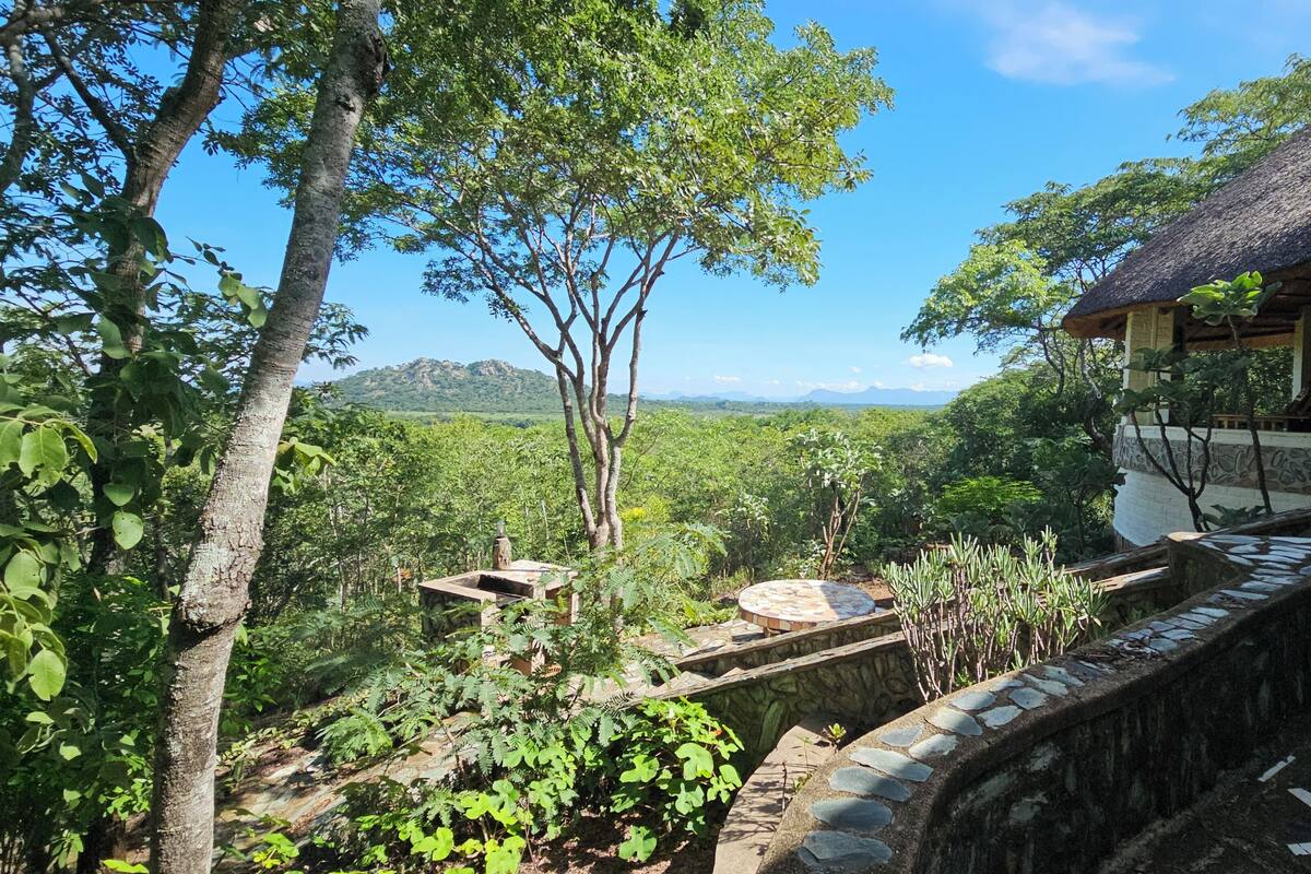 A rustic building with a thatched roof, surrounded by lush greenery and a view of distant mountains.