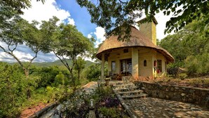 A rustic house with a thatched roof, surrounded by lush greenery and a stone pathway leading to the entrance.