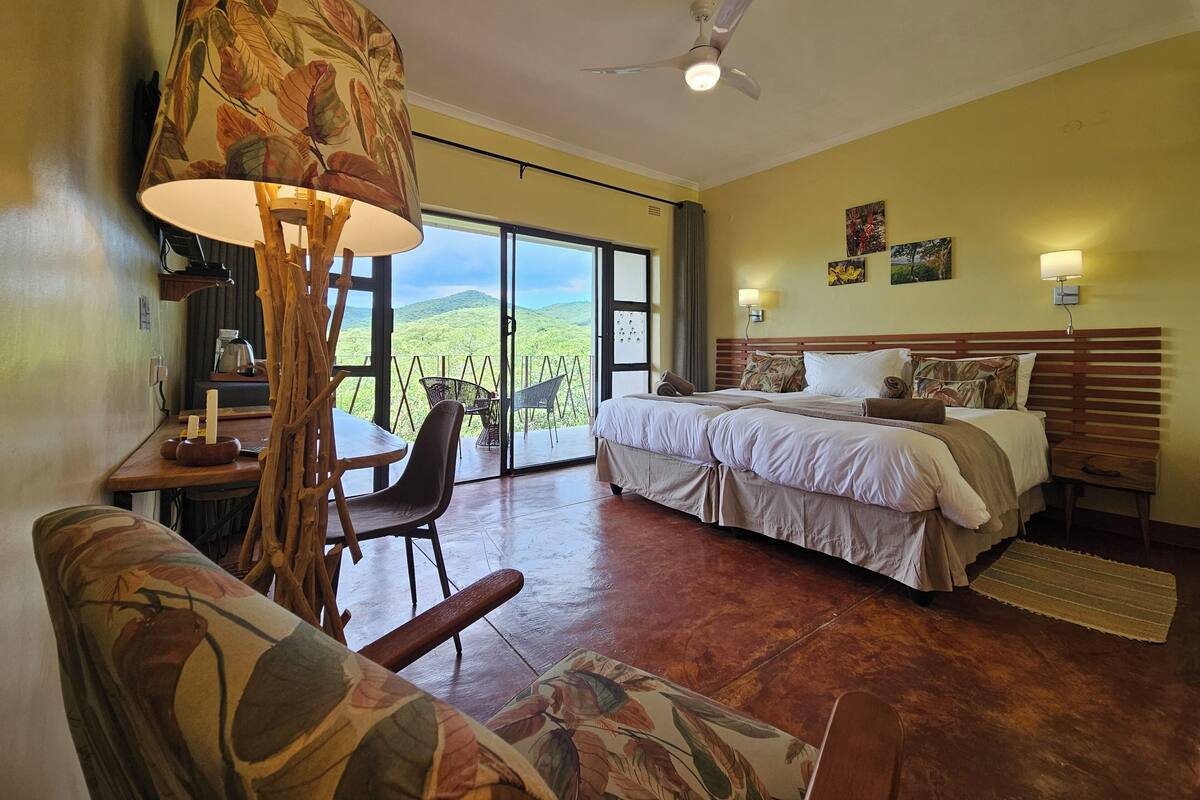 A hotel room with a bed, a desk with a lamp, a chair, and a view of the outdoors.