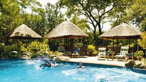 A pool area with three thatched umbrellas, lounge chairs, and a stone wall.