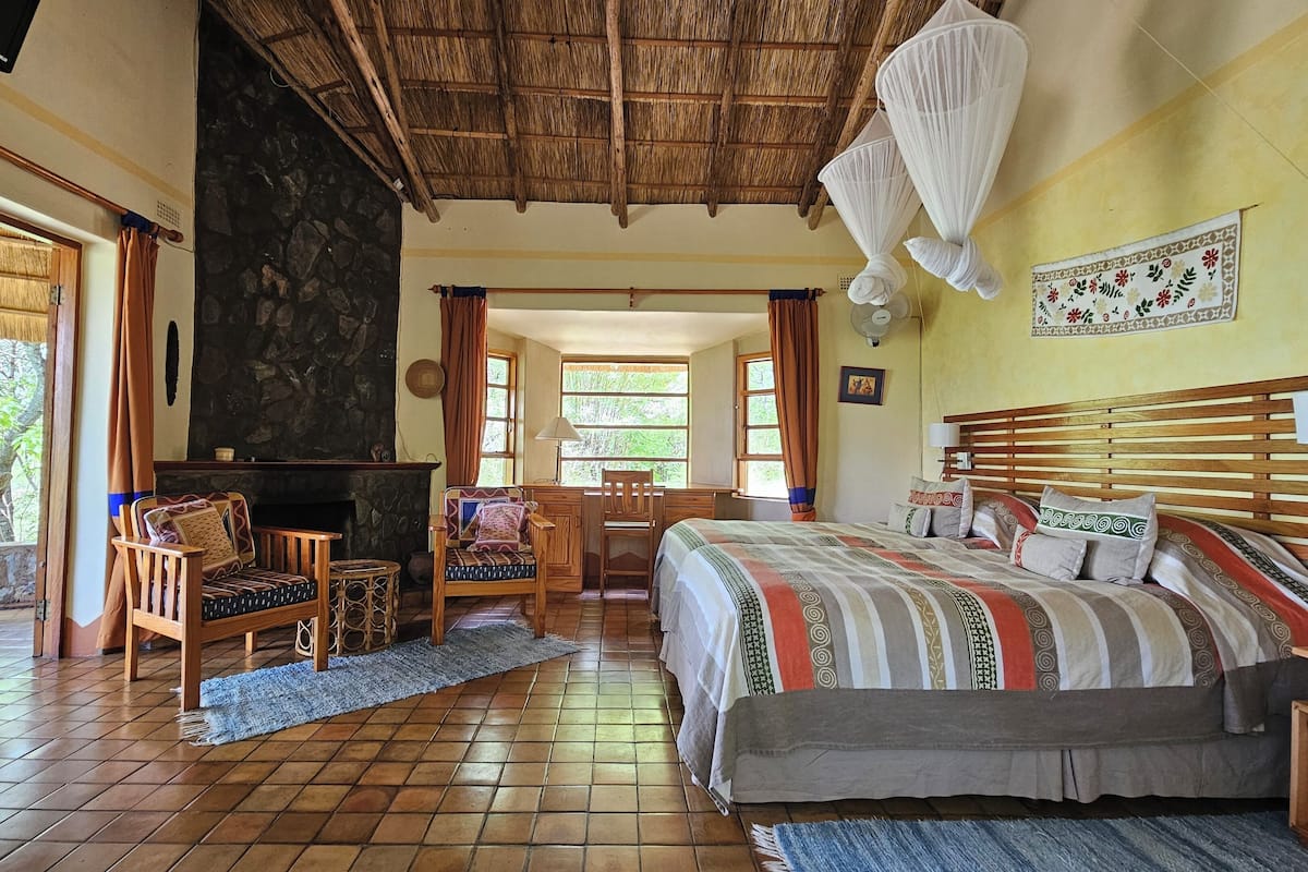 A spacious room with a large bed, a wooden desk, a fireplace, and a view of greenery outside.