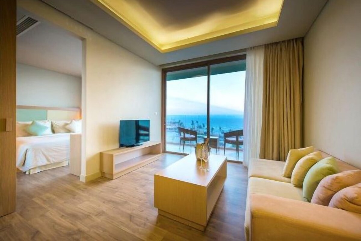 Una habitación de hotel moderna con cama, sofá, mesa de centro y televisión. La habitación tiene un ventanal con vista al mar.