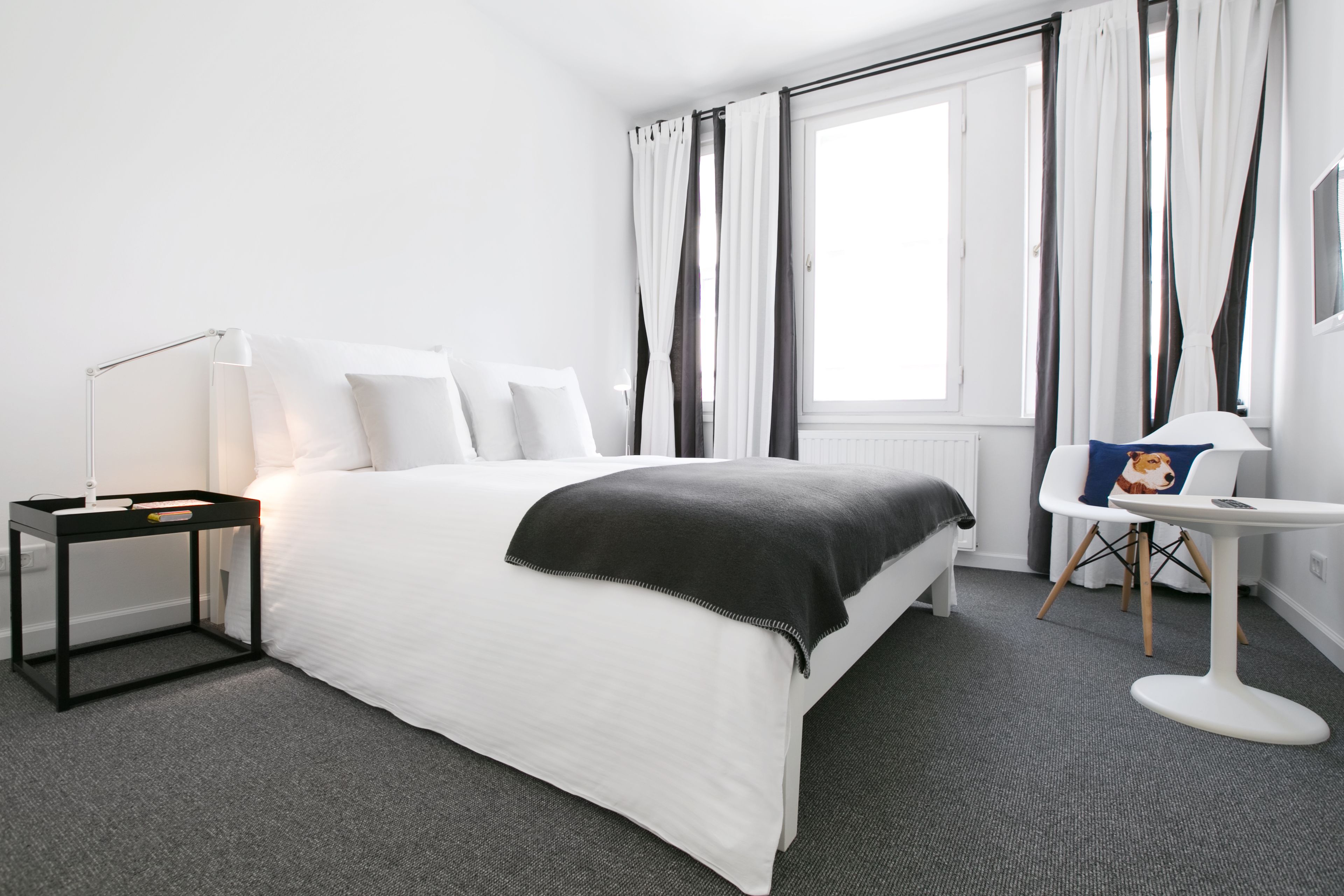Une chambre d’hôtel moderne avec un lit bien fait, une table de chevet avec une lampe, une petite table ronde avec une chaise et une fenêtre avec des rideaux.