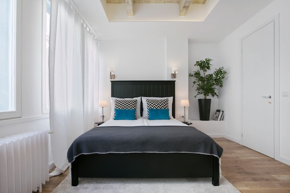 Un dormitorio moderno con una cama grande, cortinas blancas, una planta en maceta y un radiador.