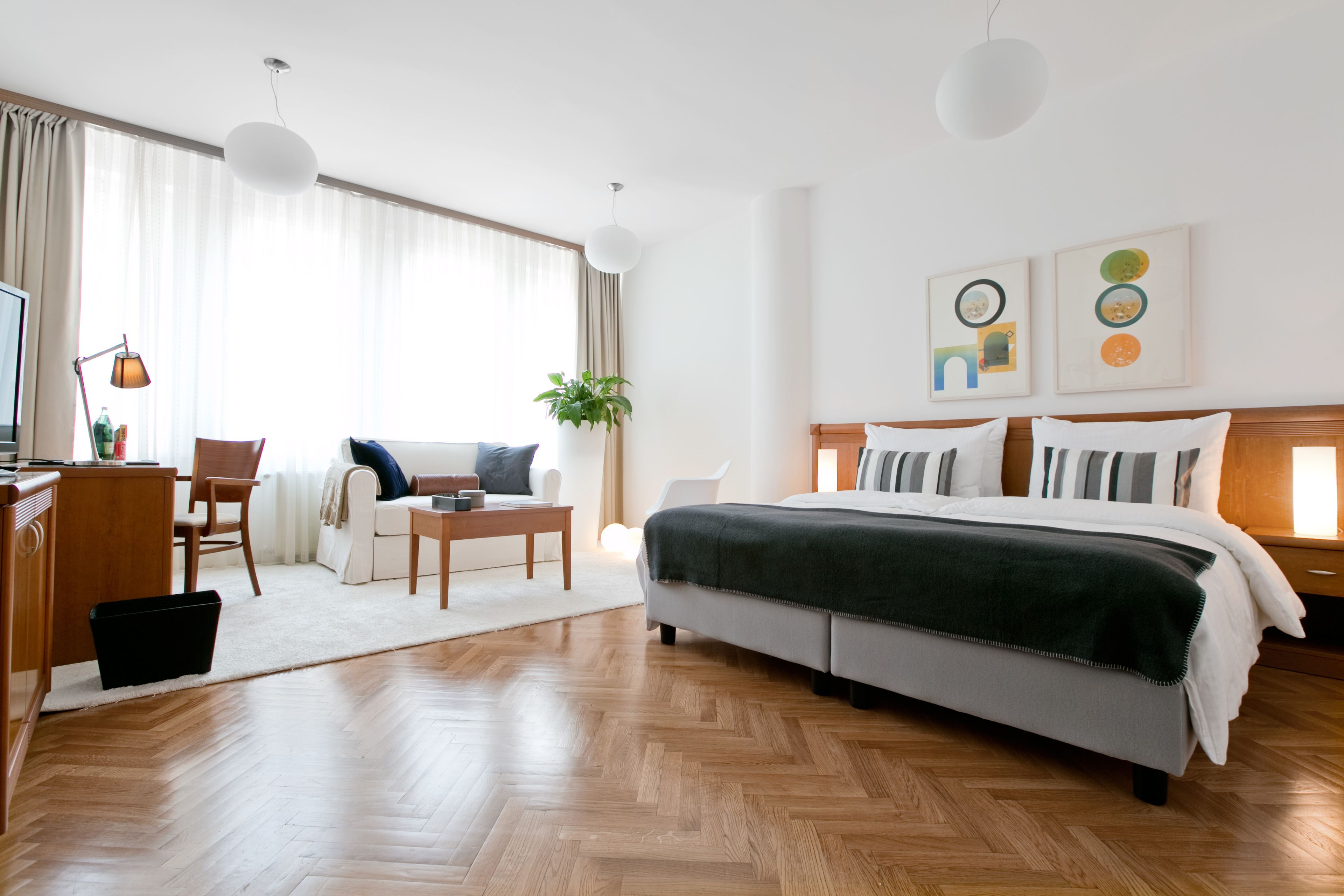 A modern hotel room with a large bed, a desk with a computer, a chair, and a sofa. There are two pendant lights and a plant on the windowsill.