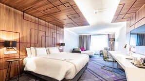 A modern hotel room with a large bed, a desk, a chair, and a window with curtains.