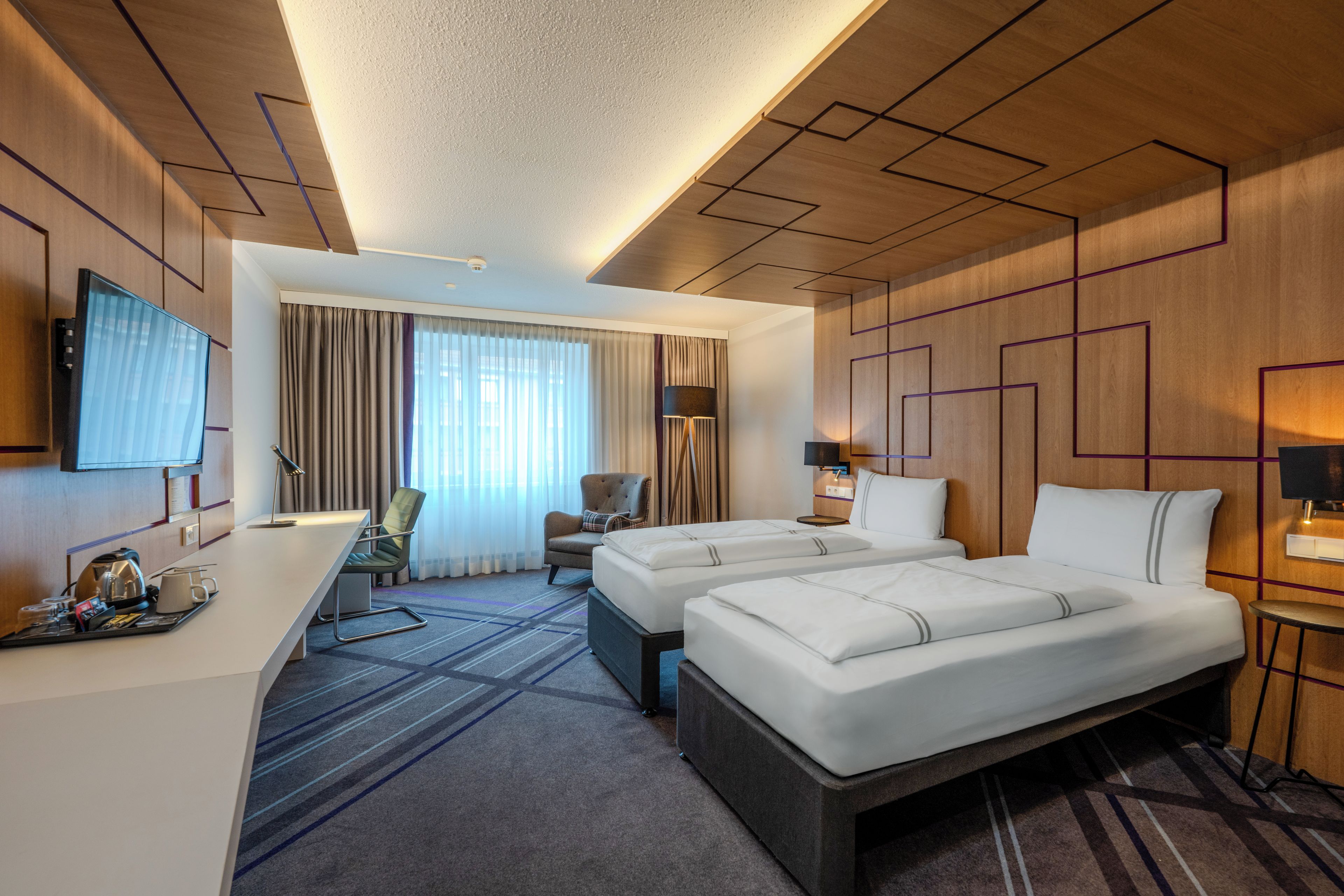 Ein Hotelzimmer mit Schreibtisch, zwei Betten, einem Sessel, einem Fernseher und einem Fenster mit Vorhängen.