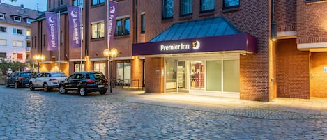 Un bâtiment en briques avec une enseigne lumineuse « Premier Inn », une allée pavée et des voitures garées.