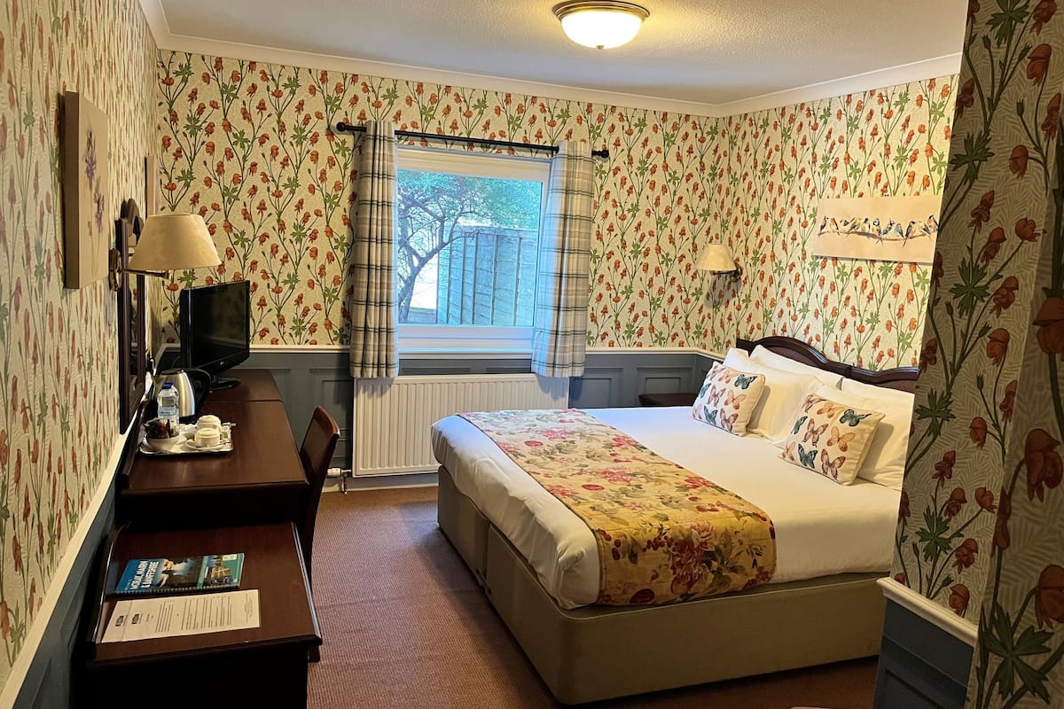 Quarto de hotel com papel de parede floral, uma cama com roupa de cama florida, uma escrivaninha com abajur e uma janela com cortinas.