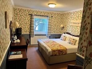 Et hotelværelse med blomstret tapet, en seng med blomsterbetræk, et skrivebord med en lampe og et vindue med gardiner.