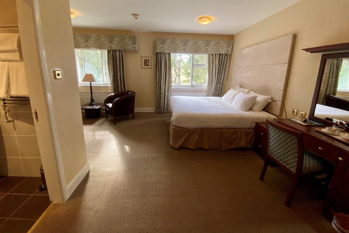 Quarto de hotel com uma cama grande, uma escrivaninha, uma cadeira, um abajur e uma janela com cortinas.