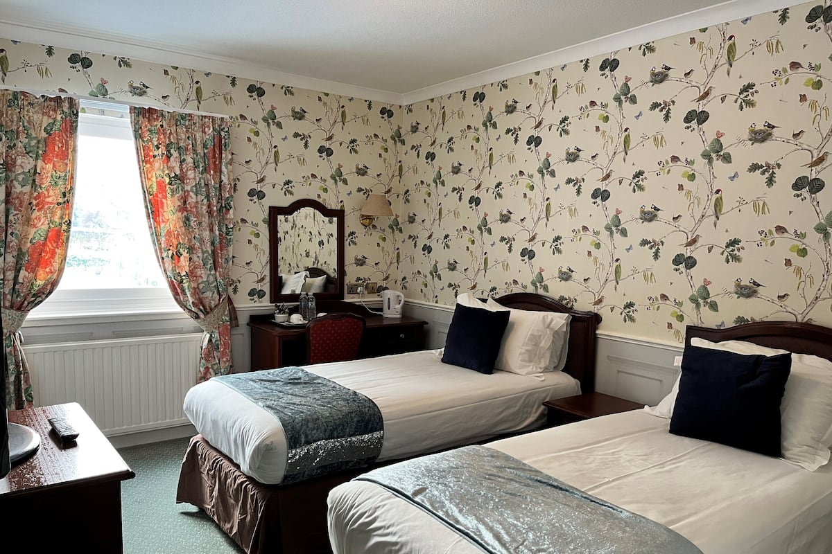 Quarto de hotel com duas camas de solteiro, uma escrivaninha com cadeira, um armário e uma janela com cortinas florais.