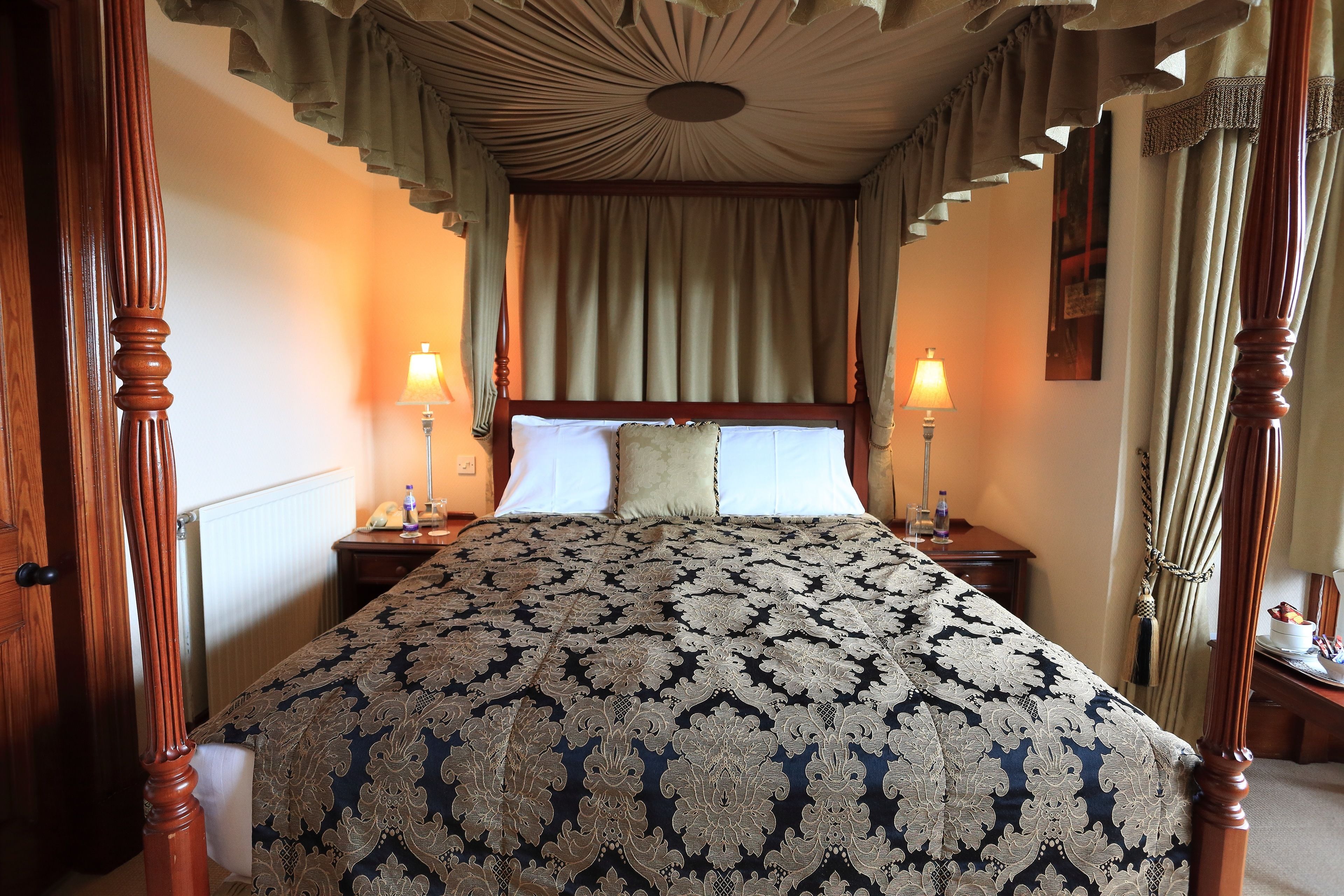 A four-poster bed with a patterned bedspread, two bedside lamps, and a nightstand with a lamp.