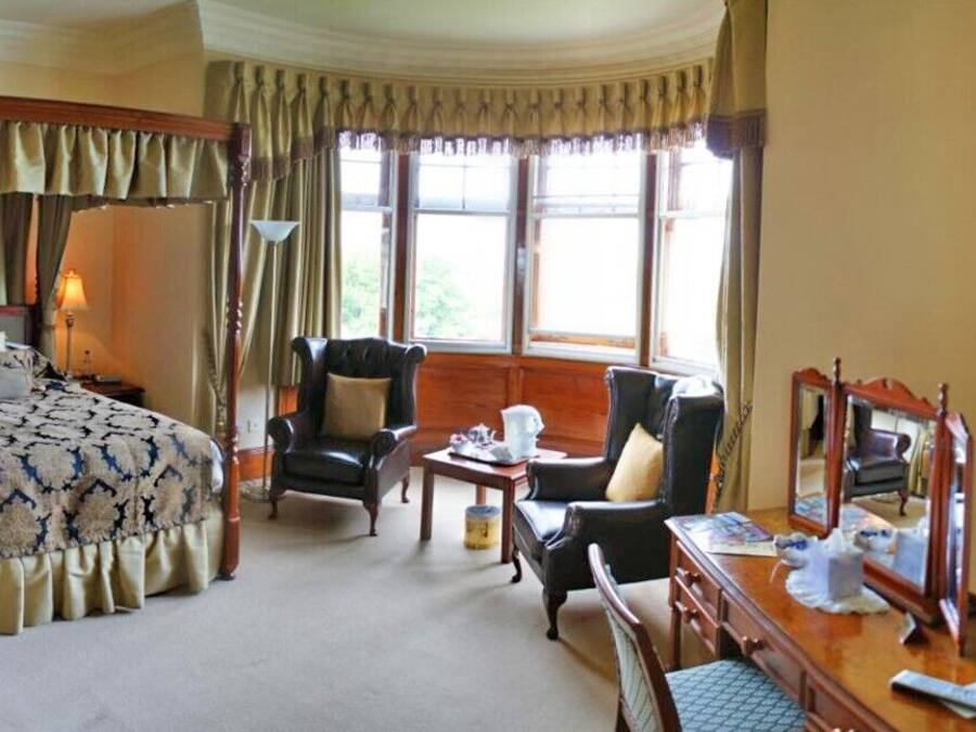 A four-poster bed with a patterned bedspread, two armchairs, a wooden desk with a mirror, and a television on the wall.