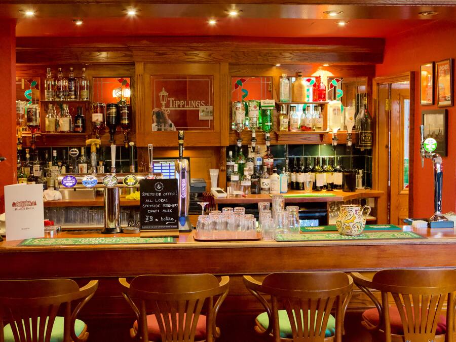 Uma área de bar com cadeiras de madeira, diversos tipos de bebidas e um aviso publicando uma cervejaria local.