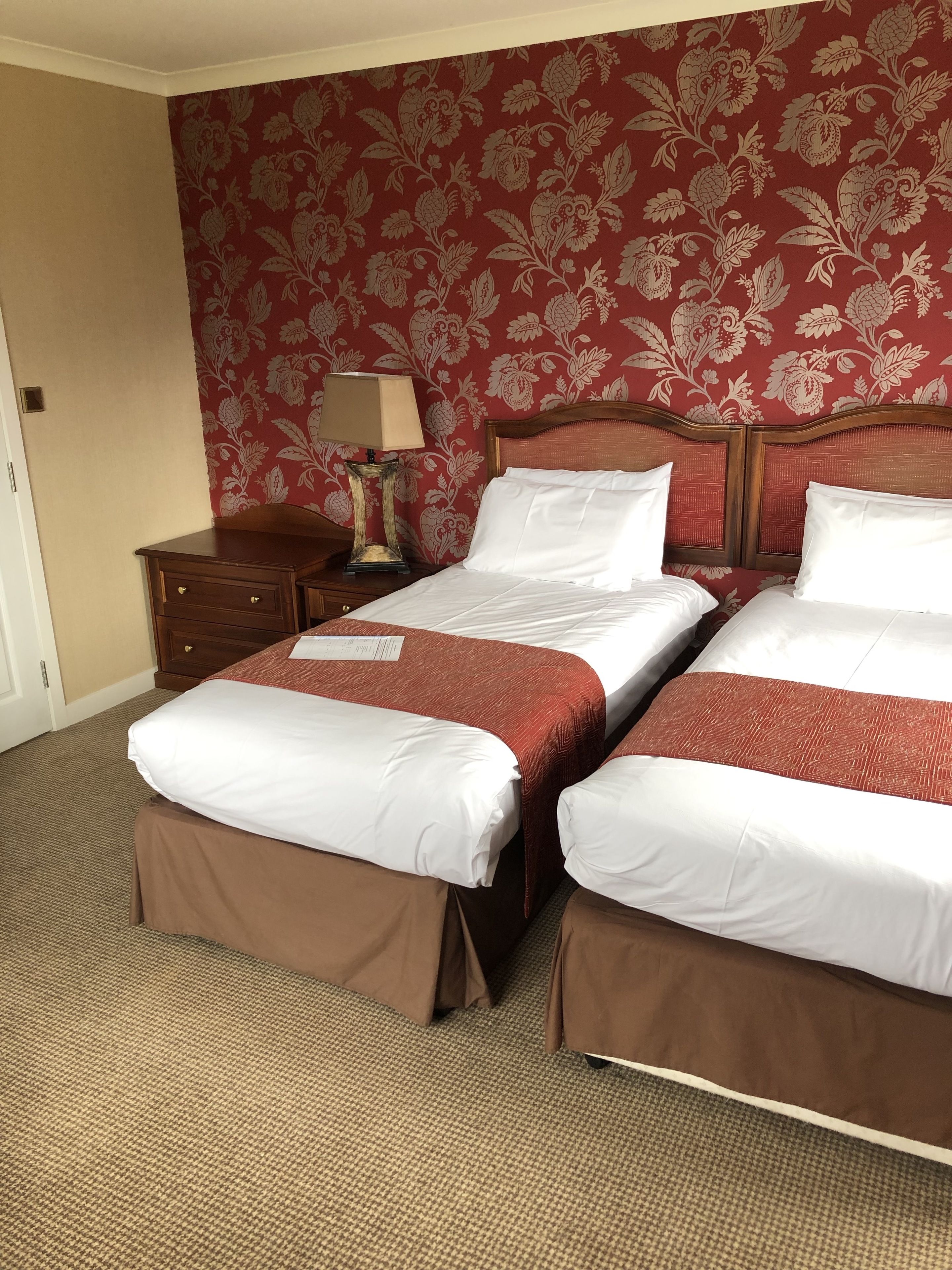 A hotel room with two beds, a wooden nightstand, a lamp, and a floral wallpaper.