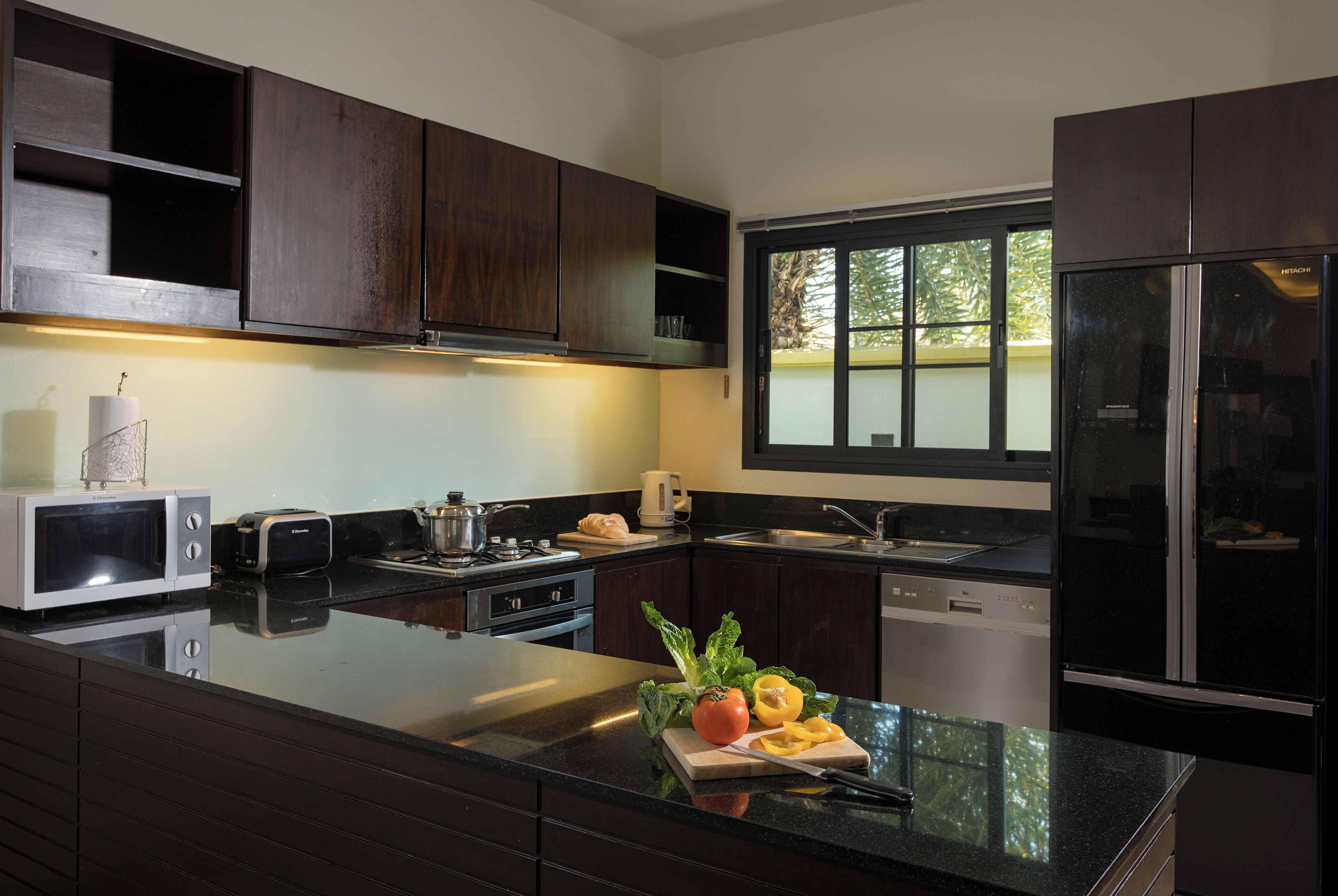 A modern kitchen with dark wood cabinets, a stainless steel refrigerator, and a large island with fresh vegetables.
